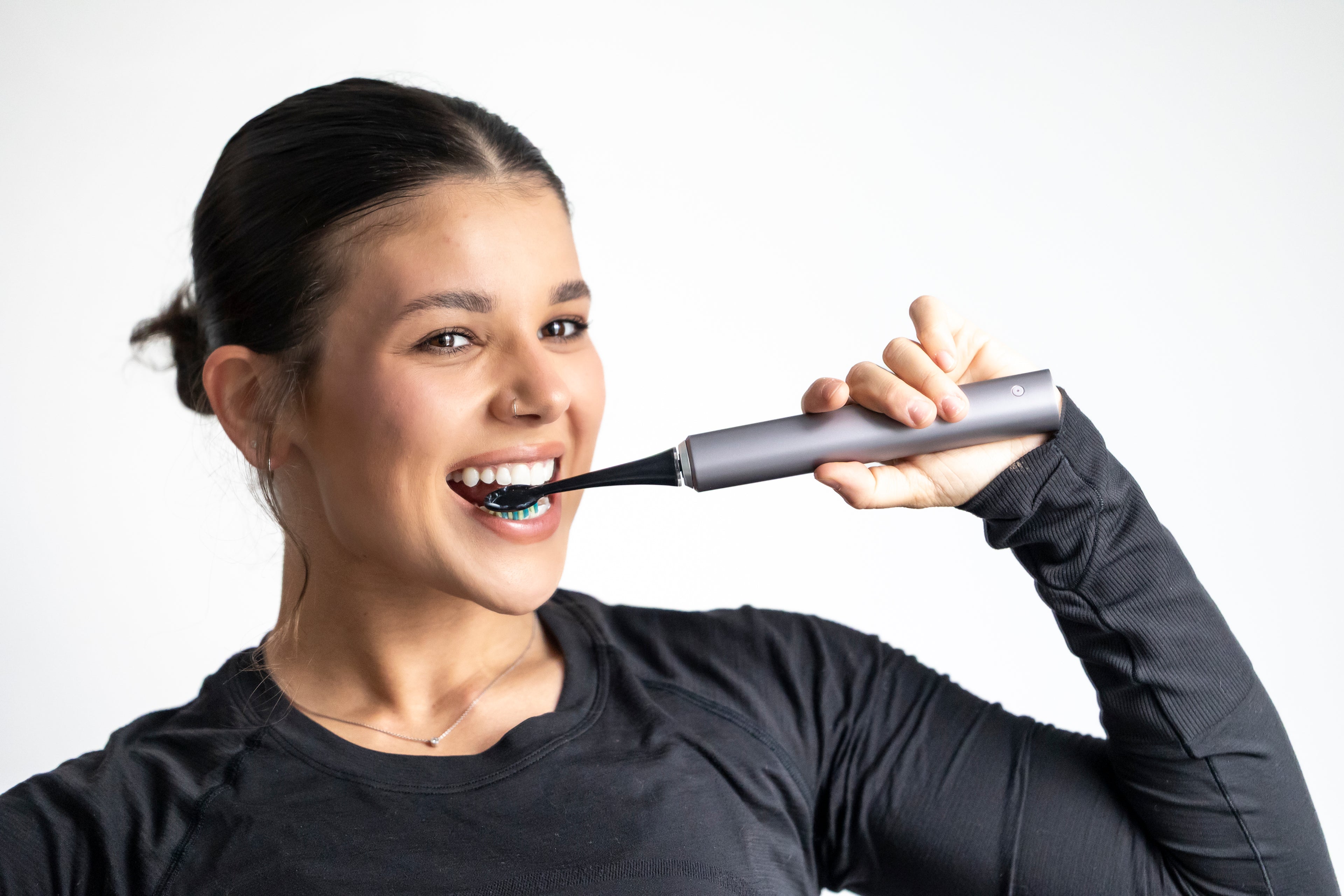 A woman with brown hair smiling with a silver electric toothbrush in her mouth.